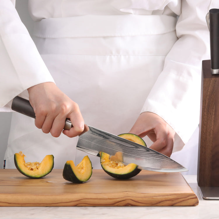 Person slicing a green and orange squash on a wooden cutting board with a Damascus steel chef knife.
