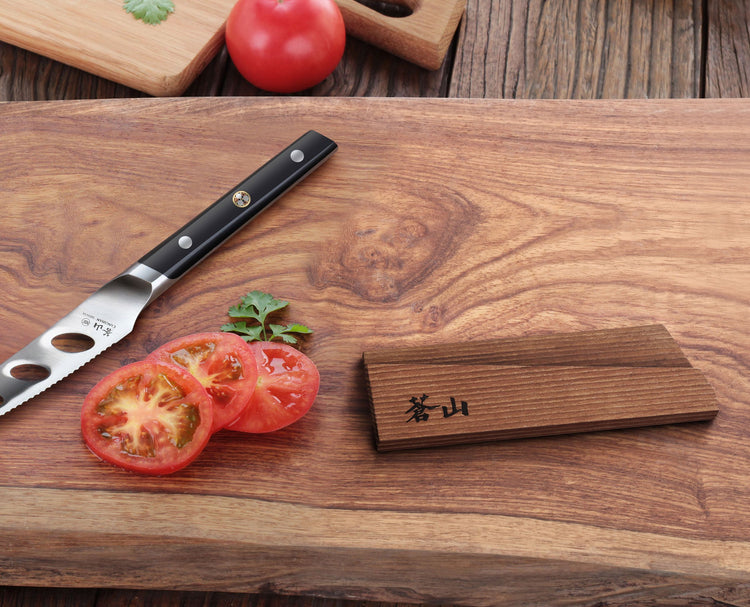 Solid ash wood magnetic knife sheath placed on a wooden cutting board next to a knife and sliced tomatoes.