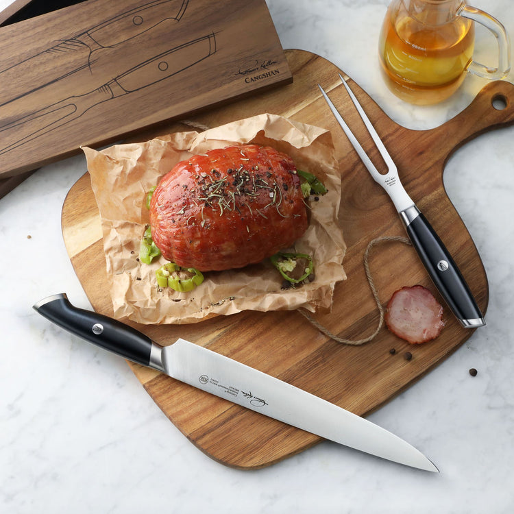 Carving knife and fork with black handles on wooden board next to cooked meat on parchment paper and glass of amber liquid.
