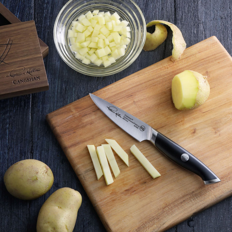 Paring knife with black handle on wooden cutting board next to peeled and cut potatoes and a bowl of diced potatoes.