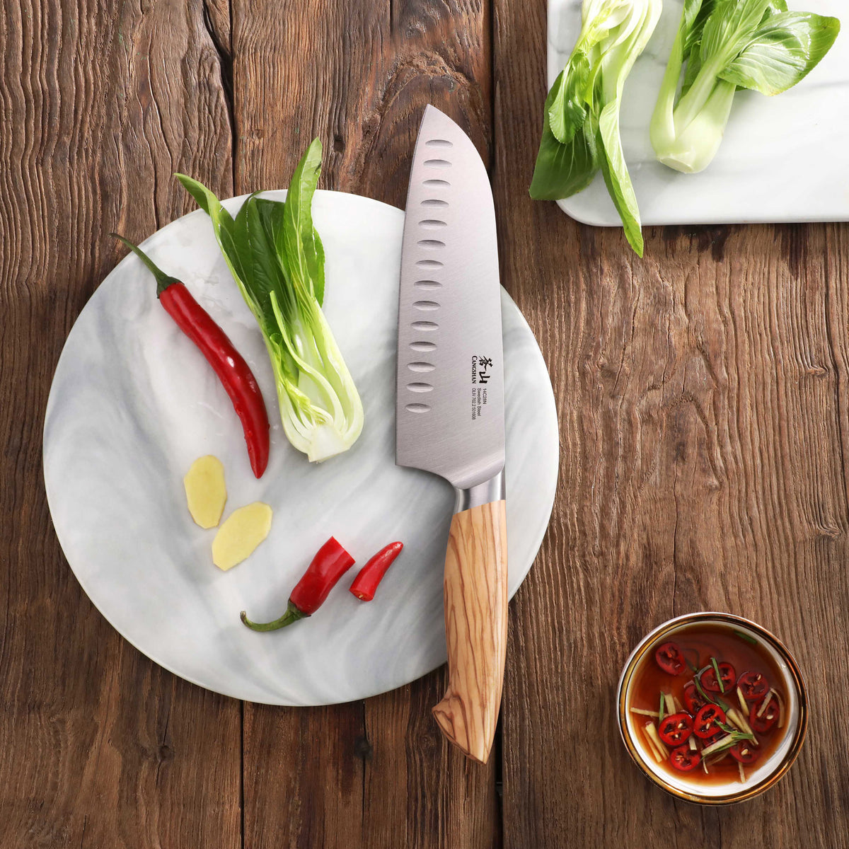 Santoku knife with wooden handle on a round marble board with chili peppers, bok choy, and ginger slices.