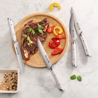 
                    
                      Four stainless steel steak knives arranged around a wooden plate with grilled steak, red and yellow bell pepper slices, and h
                    
                  