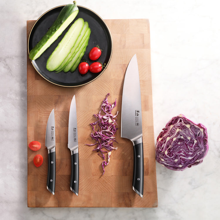 Three kitchen knives with black handles on a wooden cutting board, alongside sliced cucumber, cherry tomatoes, and shredded p