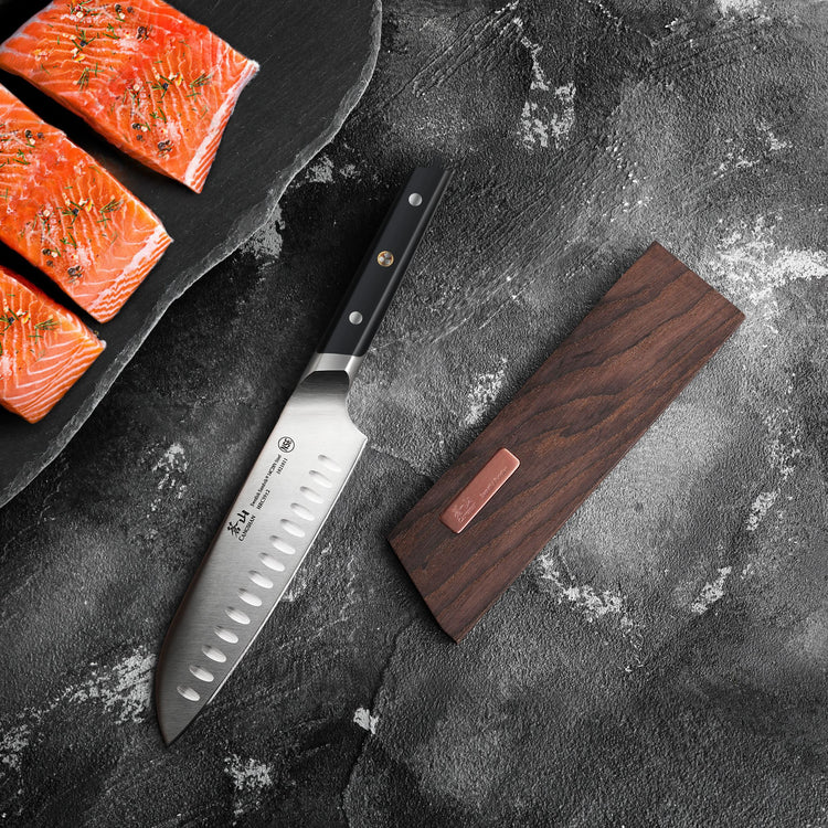 Santoku knife with black handle next to a solid wood magnetic knife sheath on a dark textured surface.