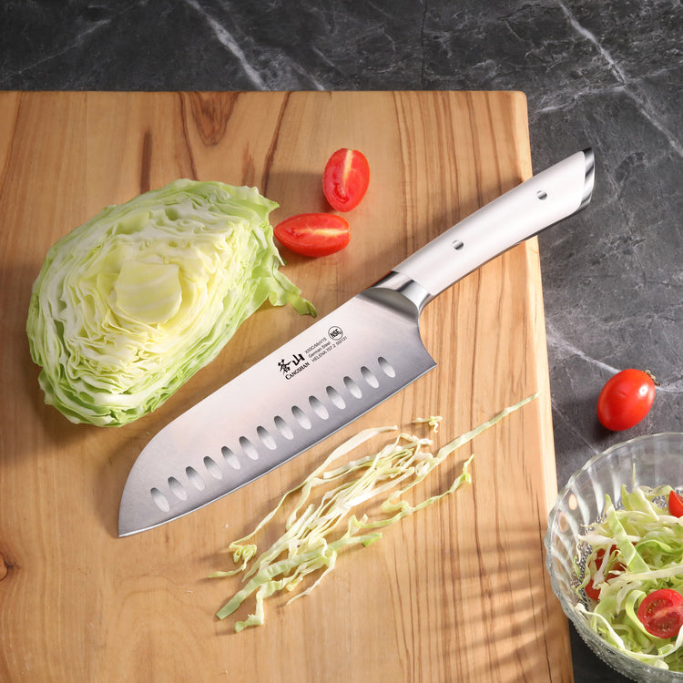 Santoku knife with a white handle on a wooden cutting board next to sliced cabbage and cherry tomatoes.