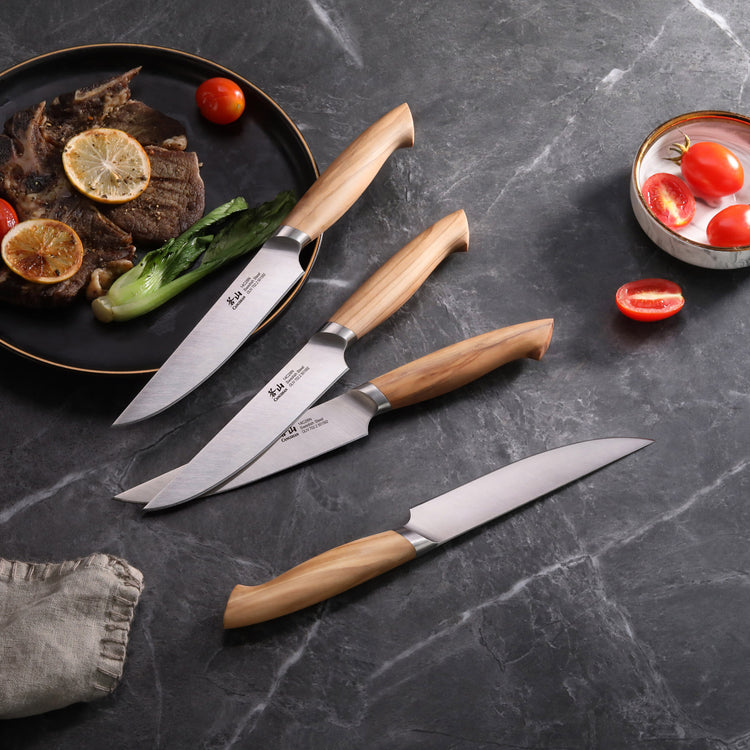 Four steak knives with wooden handles arranged on a dark surface next to a plate with cooked meat, lemon slices, and vegetabl
