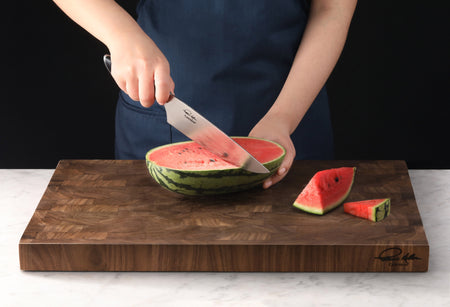 Person slicing a watermelon on a thick wooden end-grain cutting board with a chef's knife.