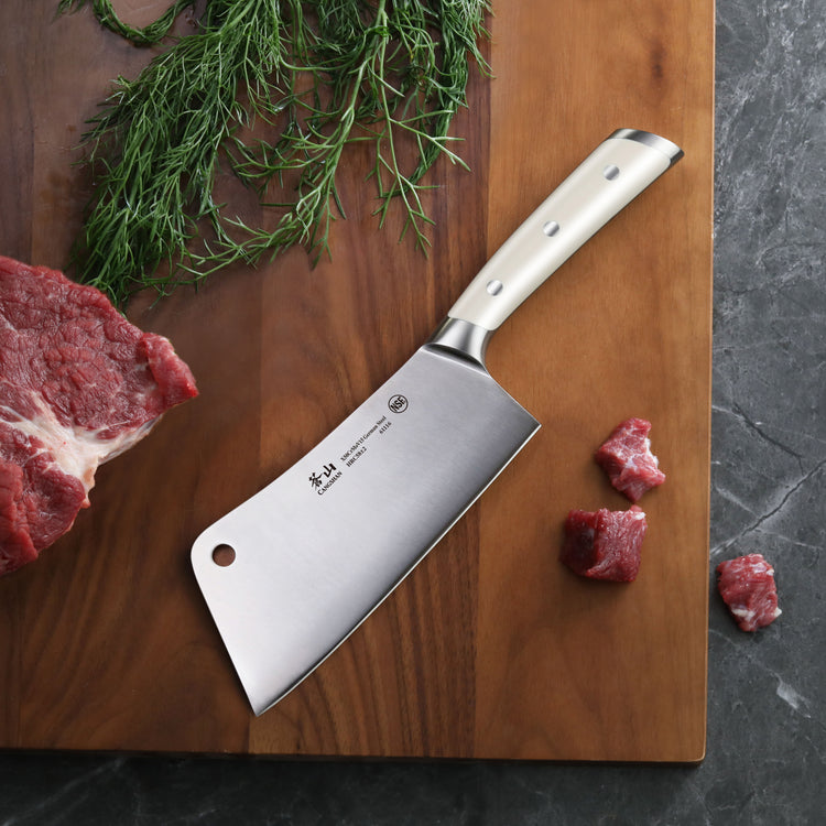 Cleaver knife with stainless steel blade and white handle on wooden cutting board with herbs and raw meat pieces.