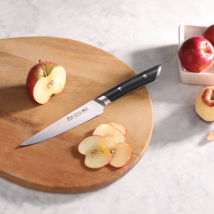 Serrated utility knife with black handle on wooden cutting board next to sliced and halved red apples.