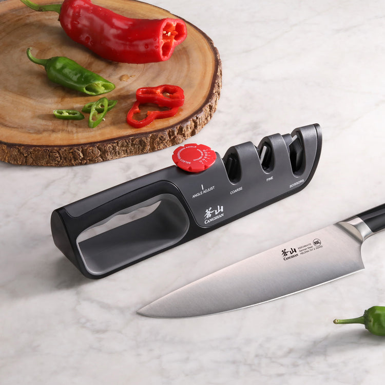 Knife sharpener with three slots and adjustable dial next to a chef's knife and sliced red and green peppers on a wooden cutt