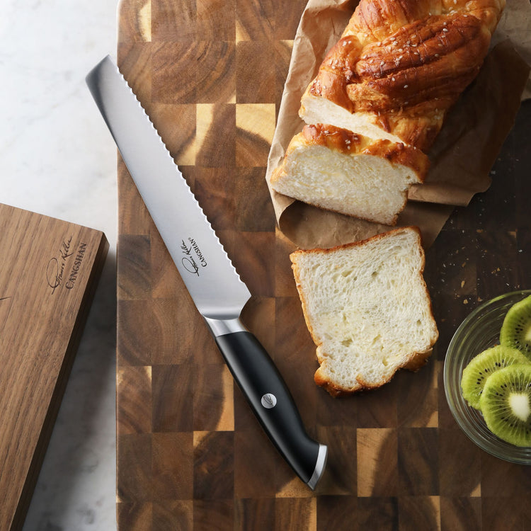 Serrated bread knife with black handle on wooden cutting board next to sliced loaf and kiwi fruit bowl.