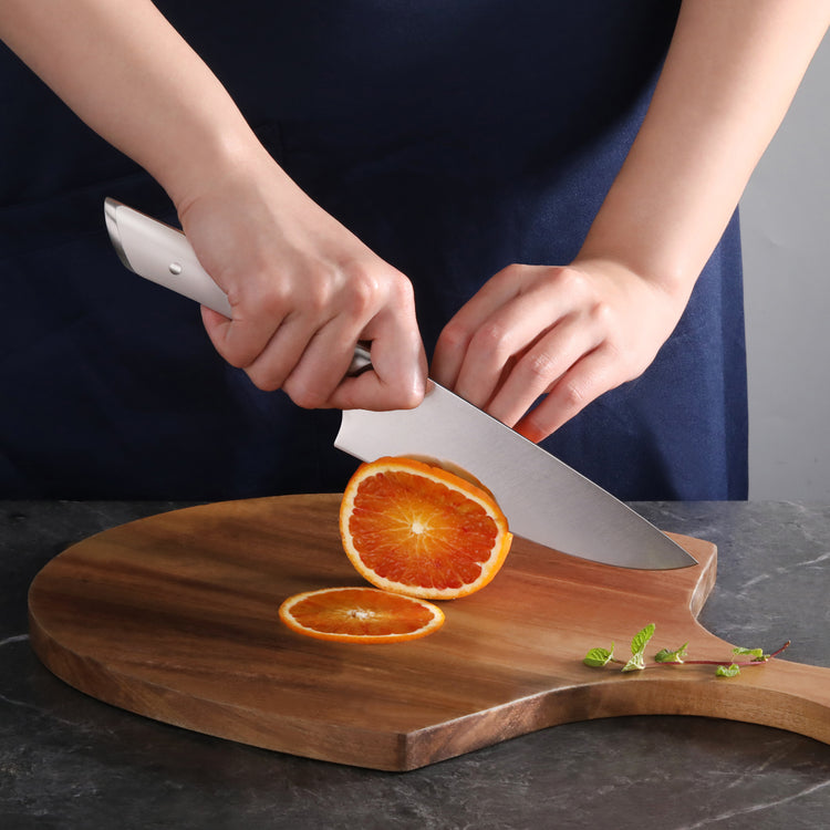 Hands slicing an orange on a wooden cutting board with a chef's knife.