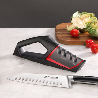 
                    
                      Black and red handheld knife sharpener with four sharpening slots placed on a marble surface next to a chef's knife and a cut
                    
                  