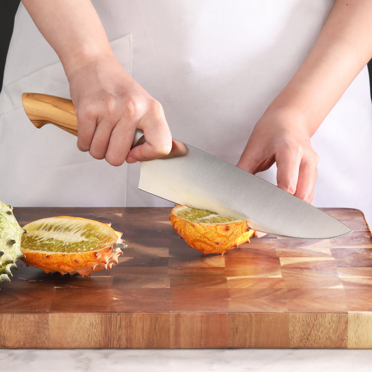 Person using an 8-inch chef's knife with an olivewood handle to cut a kiwano fruit on a wooden cutting board.