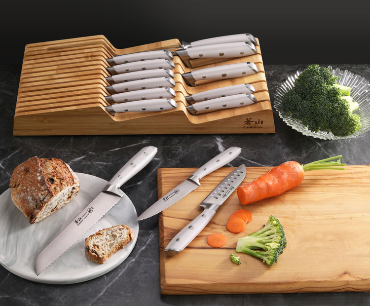 15-piece knife set with white handles in bamboo tray, two knives cutting bread and vegetables on marble countertop.