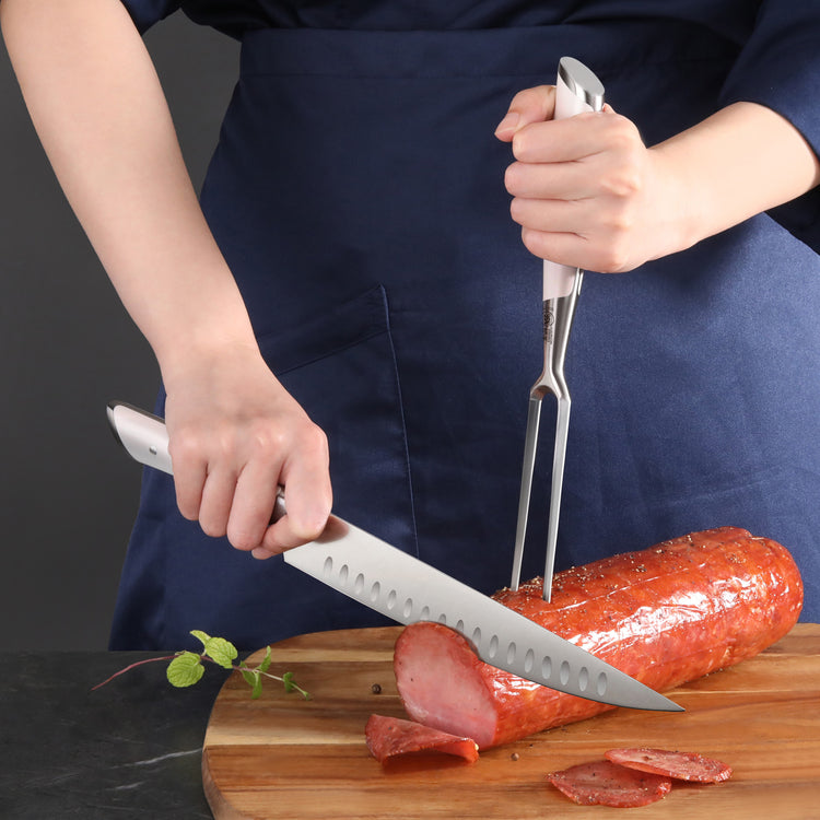 Person slicing a large cooked sausage on a wooden board using a carving knife and fork with white handles.