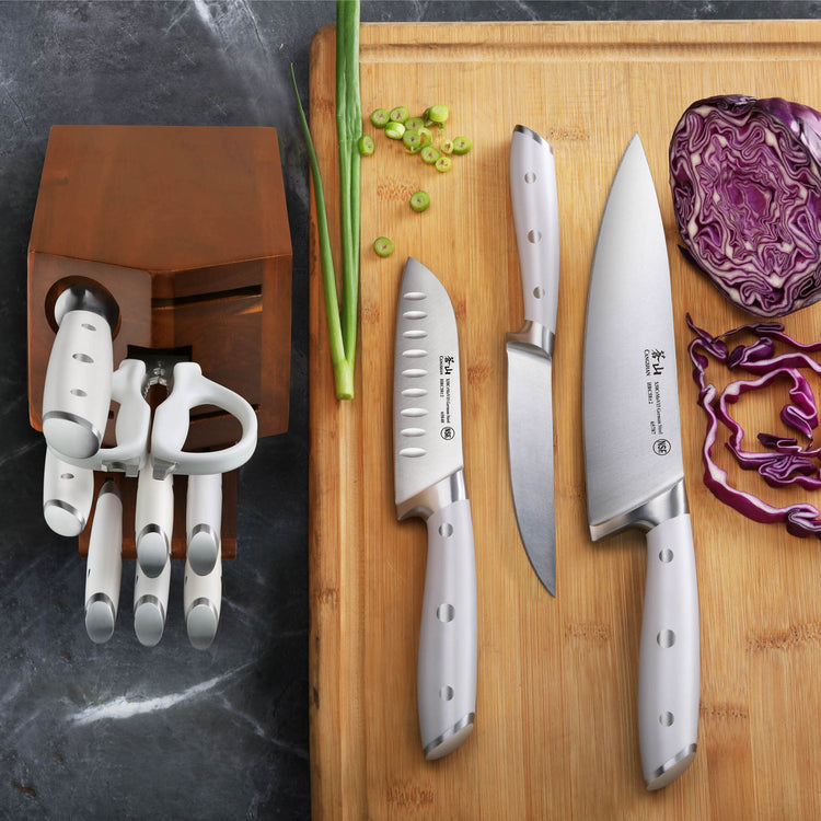 Set of three stainless steel knives with white handles on a wooden cutting board next to chopped green onions and purple cabb