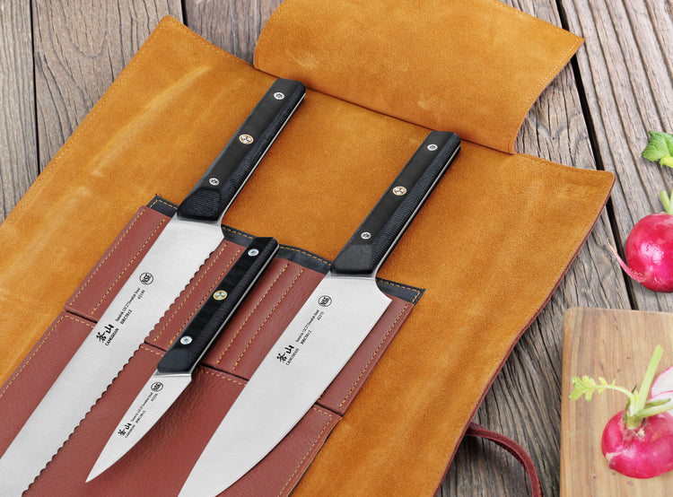 Four-piece knife set with black handles in an open tan leather roll on a wooden surface.
