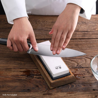 
                    
                      Hands sharpening a large kitchen knife on a whetstone with a wooden base on a wooden surface.
                    
                  