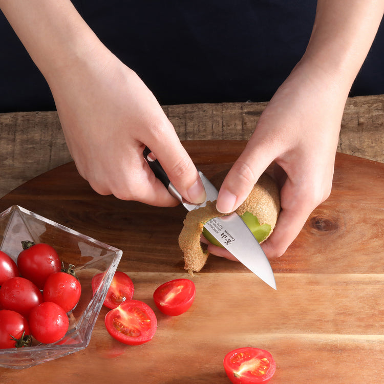 Hands peeling a kiwi with a small black-handled paring knife on a wooden cutting board next to halved cherry tomatoes.