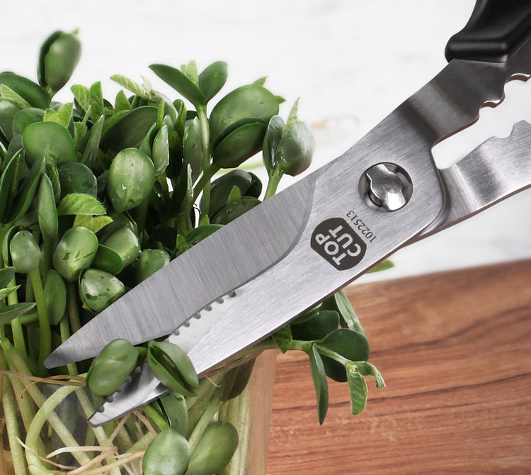 Stainless steel kitchen shears cutting green plant stems on a wooden surface.