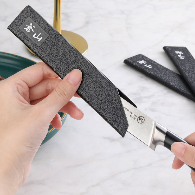 Hands placing a black textured knife guard onto a steak knife blade on a marble surface.