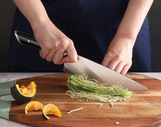 
                    
                      Person slicing shredded cabbage on a wooden cutting board with a large kitchen knife, next to cut pieces of squash.
                    
                  