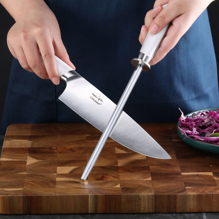 Hands holding a knife and a white-handled honing steel on a wooden cutting board next to a bowl of shredded purple cabbage.