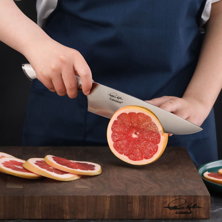 Hand holding a knife cutting a grapefruit on a wooden cutting board with sliced grapefruit pieces nearby.