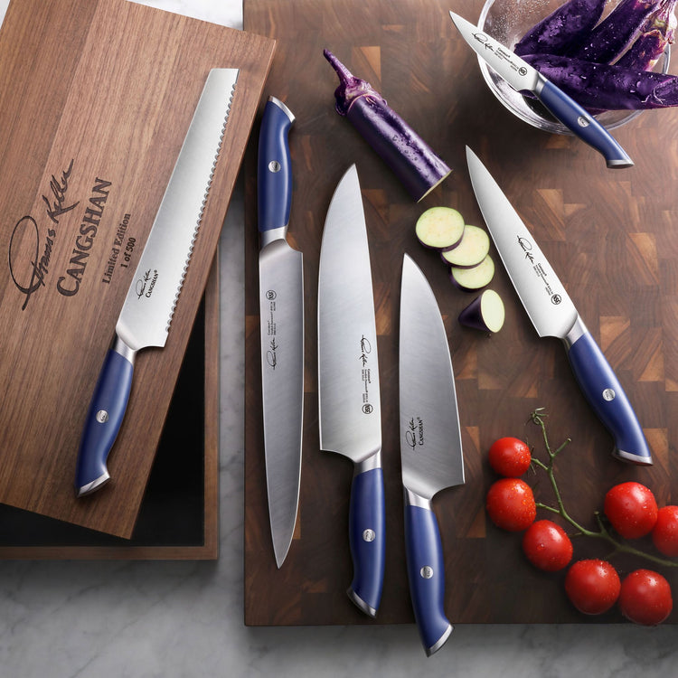 Six blue-handled kitchen knives arranged on a wooden cutting board with sliced vegetables and a wooden knife storage box.