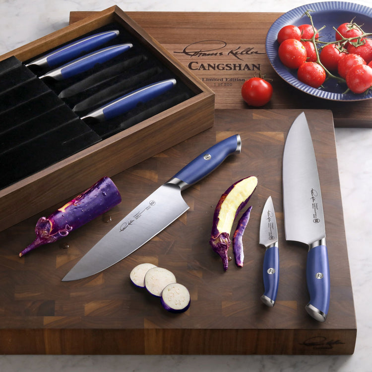 Six-piece blue-handled knife set displayed on a wooden cutting board with sliced eggplant and a plate of tomatoes.