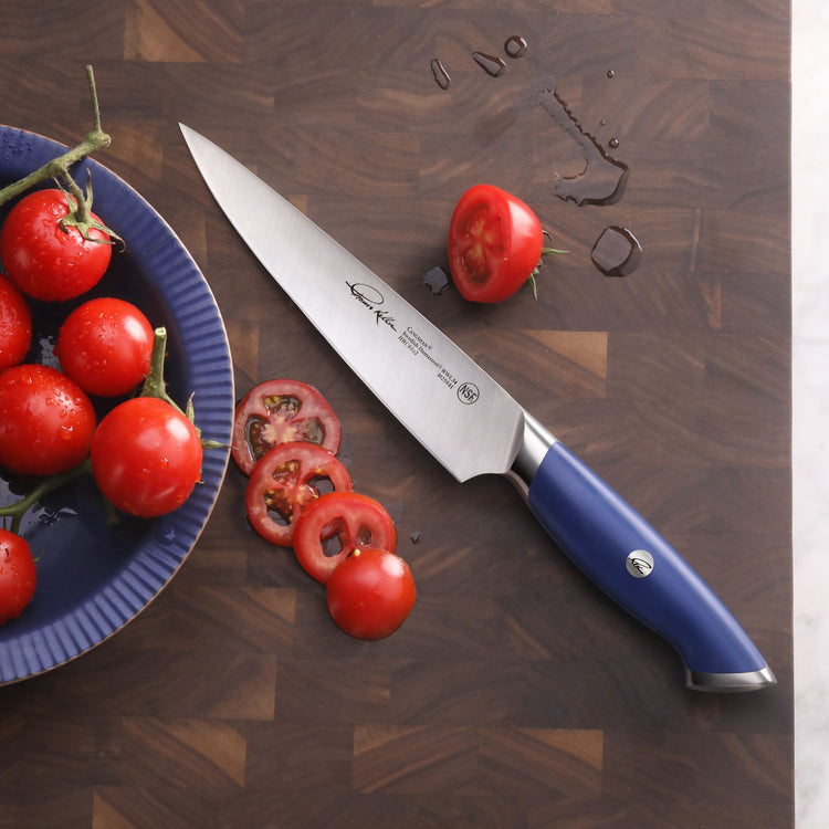 Blue-handled chef knife with sliced tomatoes on a wooden cutting board next to a blue plate of whole tomatoes.